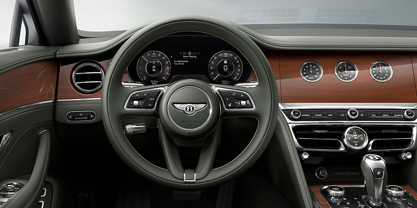 Bentley St Peter New Bentley Flying Spur sedan front cabin in Cumbrian Green hide and Crown Cut walnut veneer showing steering wheel and Rotating Display with three clocks on face