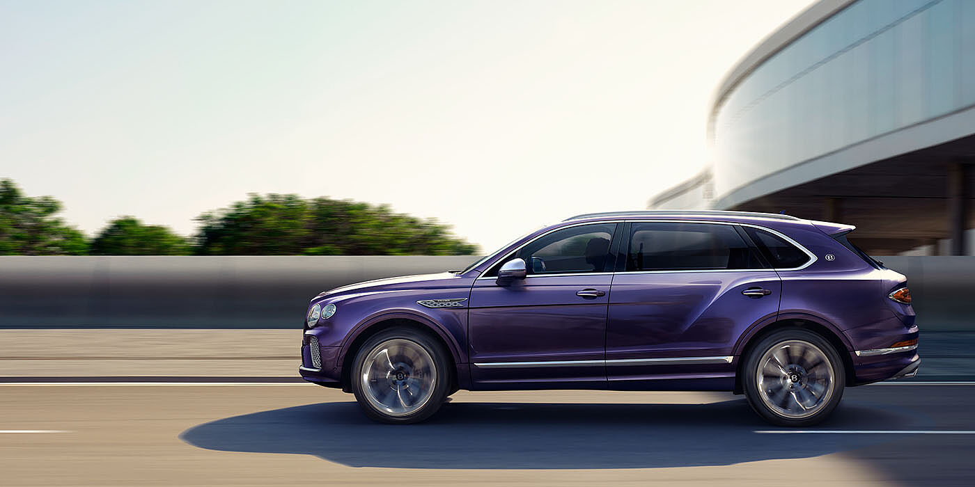 Bentley St Peter Bentley Bentayga Extended Wheelbase Mulliner luxury SUV profile in Tanzanite Purple paint, driving on an country road past trees.