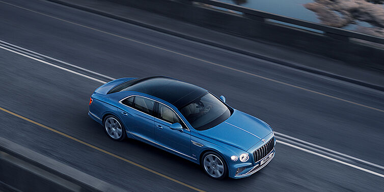 Bentley St Peter New Bentley Flying Spur Azure sedan in Light windsor blue paint by Mulliner, with 22 inch Azure wheels seen from overhead driving dynamically on a country road