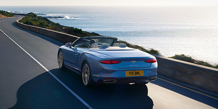 Bentley St Peter New Bentley Continental GTC Azure convertible rear view in Light Windsor blue paint with 22 inch ten spoke wheels driving dynamically on an ocean road in the sunshine